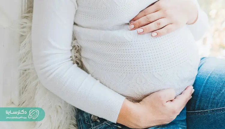 Pregnant woman suffering from vaginal varicose veins during pregnancy sitting on a sofa with her hands on her stomach wearing jeans and a white dress