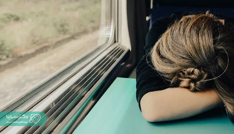 A tired woman resting her head on a table near a window, possibly experiencing fatigue related to anemia, which may be detected through an MCH test