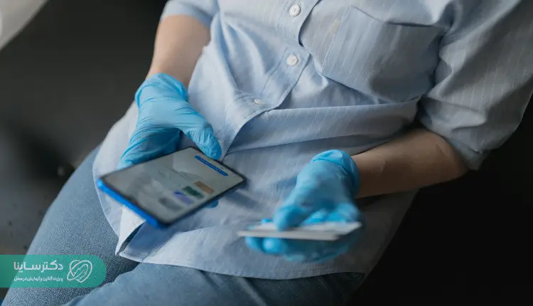 A person wearing medical gloves using a smartphone and holding a card, possibly booking a home visit for an MCH test through an online health service